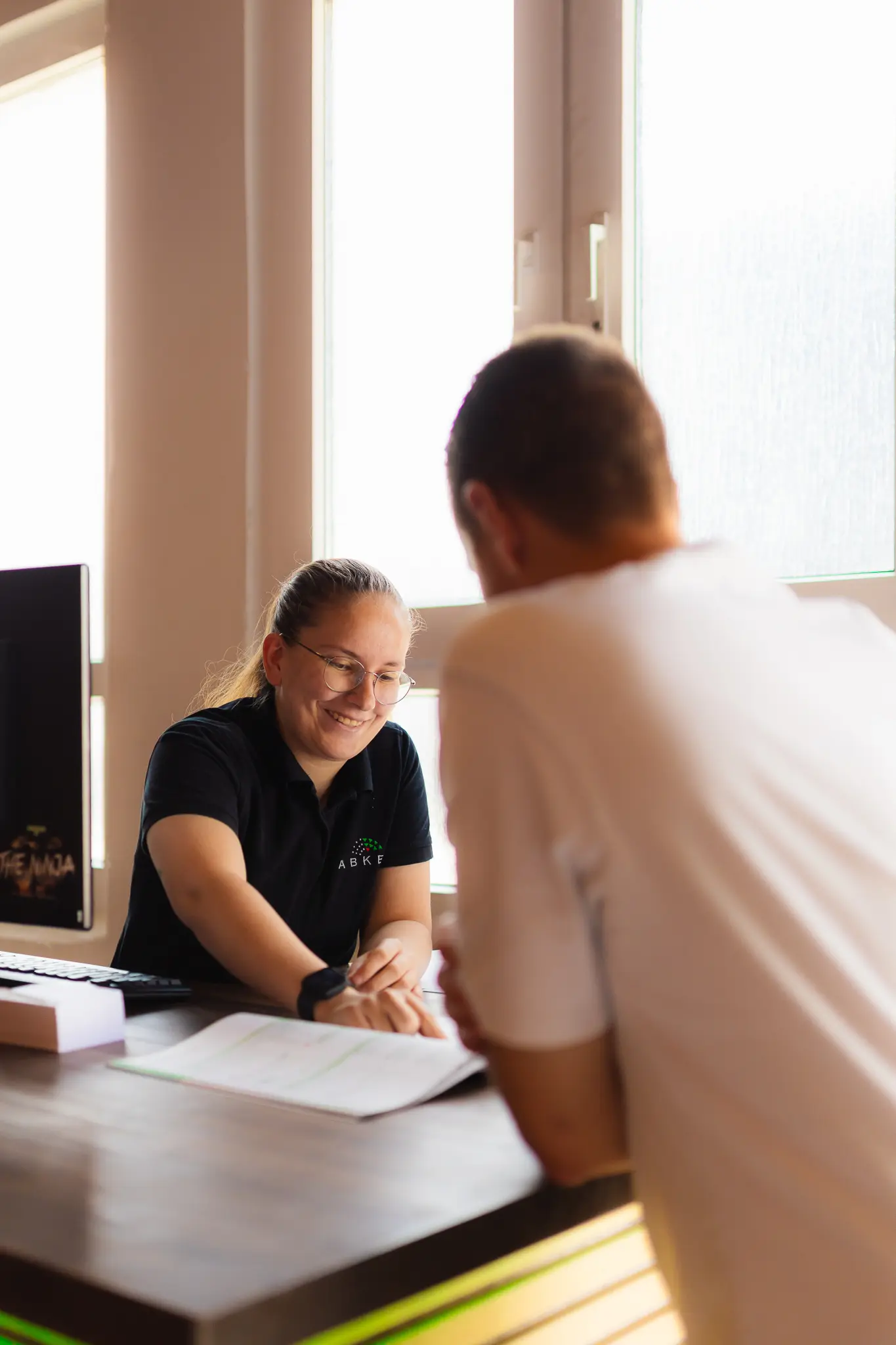 Frau mit Brille im schwarzen Poloshirt berät am Empfangstresen einen Kunden.