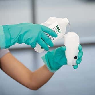 Flüssigkeit wird in den Behälter gegossen Person in grünen Handschuhen gießt Flüssigkeit aus einer Flasche in einen weißen Behälter.