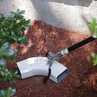 Gebogenes Gartengerät für Mulch und Erde Gartengerät mit einer gebogenen Metallschaufel, die auf Mulch in der Nähe von Sträuchern ruht.