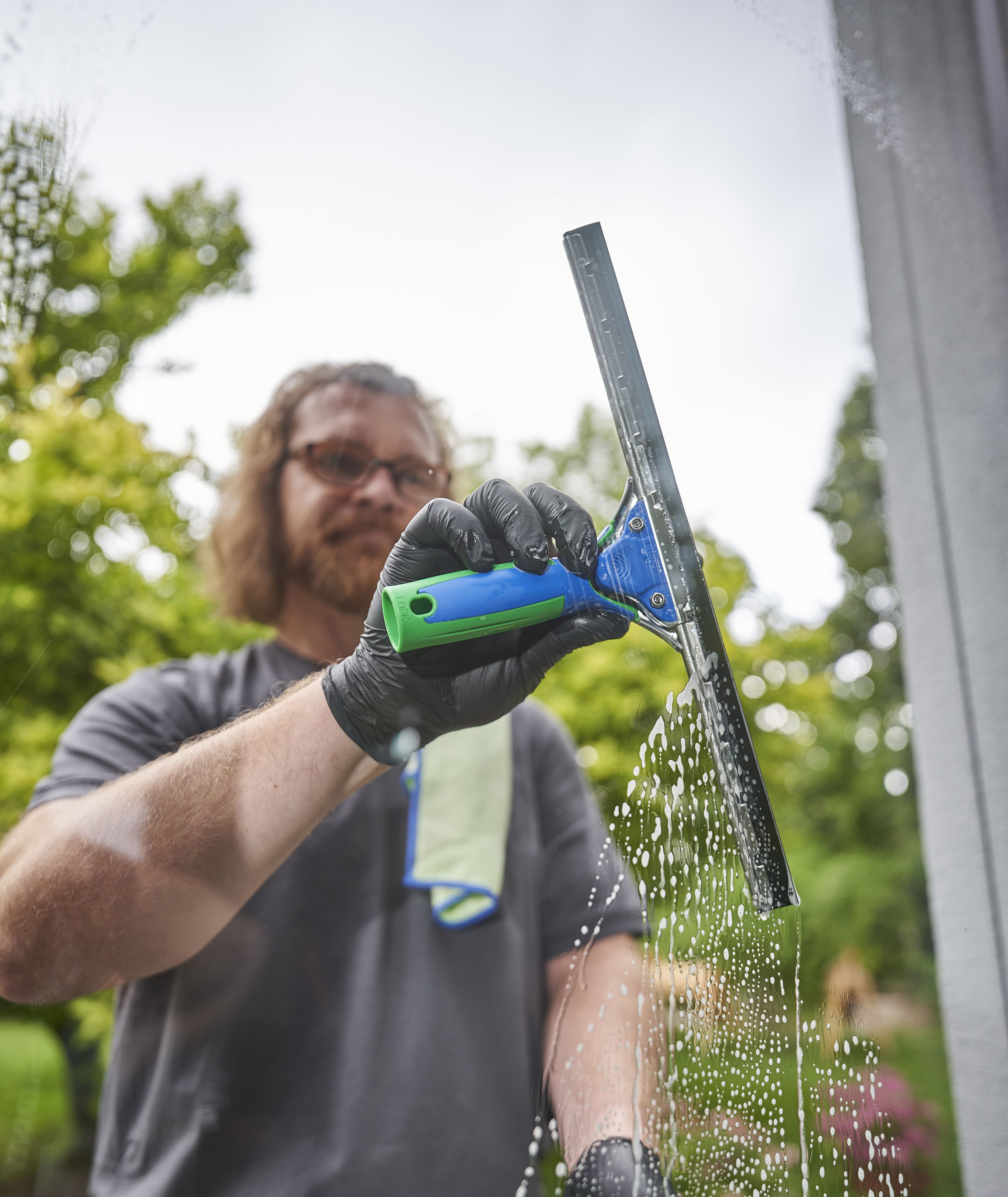 Fensterwischer mit grün-blauem Griff und schwarzer Gummilippe reinigt eine Glasscheibe mit Schaumrückständen