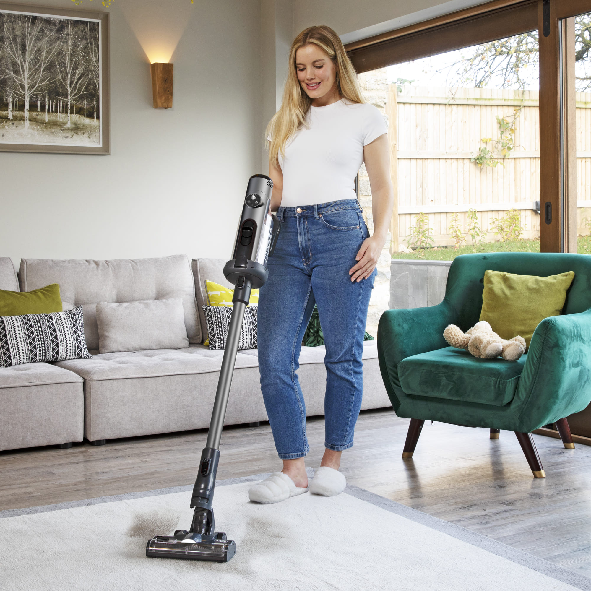 Akku-Staubsauger mit grauem und schwarzem Finish, bedient von einer Frau auf einem hellen Teppich im Wohnzimmer.