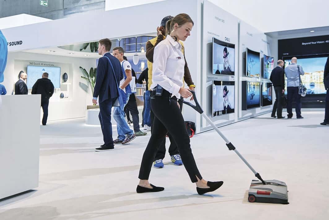 Akkubetriebene Bodenkehrmaschine für die professionelle Reinigung Frau bedient eine kompakte Akku-Kehrmaschine mit langem Stiel zur effizienten Bodenreinigung in einer Messehalle
